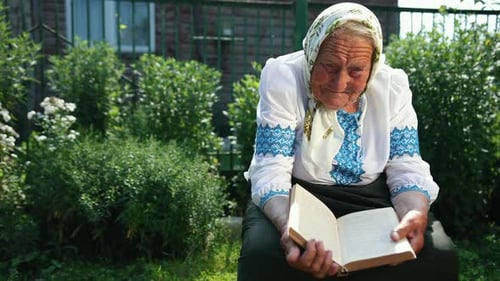 An Old Grandmother is Sitting on the Street and Reading a Book