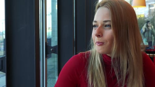 A Young Attractive Woman Sits at a Table in a Cafe, Eats Pie and Looks Out a Window with a Smile