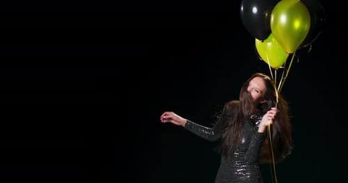A Woman in a Black Brocade Dress Dances While Holding Black and Gold Balloons Against a Black Studio