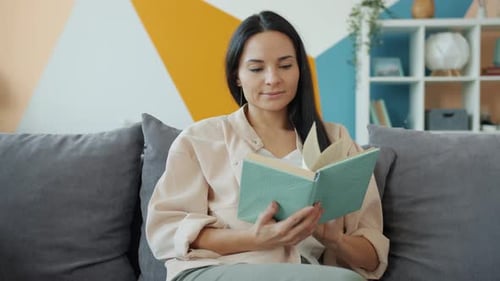 Good-looking Young Woman Reading Book and Smiling Then Turning Page in Nice Apartment