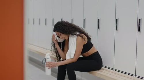 Young Active Sporty Woman Resting After Hard Workout at Gym Locker Room Drinking Water and Wiping