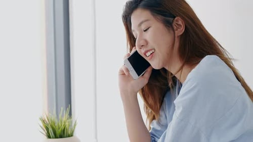 Die schöne junge Asiatin telefoniert zu Hause neben dem Fenster im Schlafzimmer.