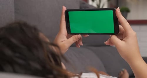 woman on the living room couch watching something on her cell phone, green screen on smart phone