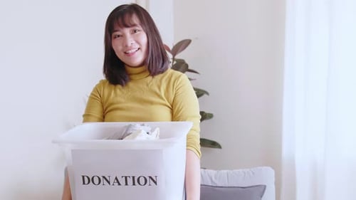 Portrait of young Asian woman looking at camera holding box with clothes for donation. Concept campa