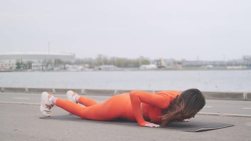 A beautiful woman in a bright orange fitness suit is doing exercises on the street. A young girl goe