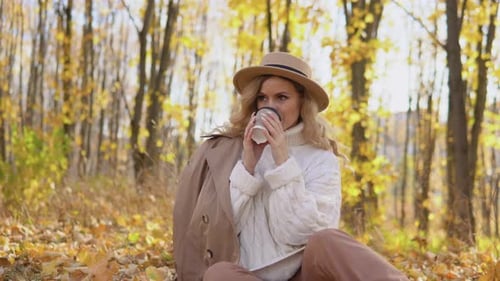 Beautiful Blonde Woman in a Brown Hat Drinks Coffee in the Autumn Park