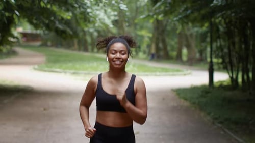 Follow Shot of Sporty African American Woman Jogger Running in Public Park Listening to Music and