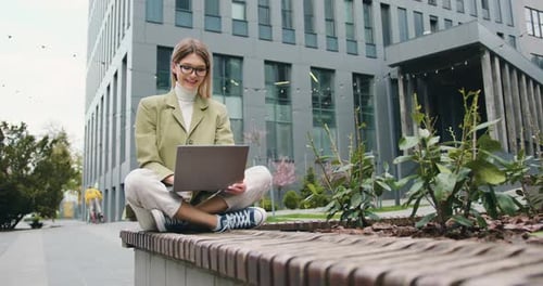 Mujer joven segura trabajando al aire libre sentada cerca del edificio de oficinas escribiendo en la computadora portátil