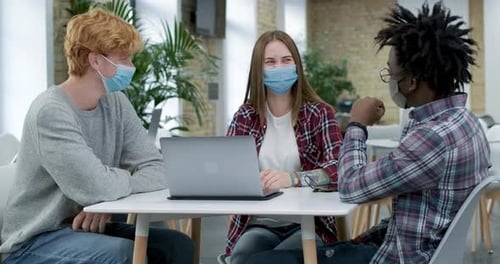 Three Millennial Coworkers in Covid-19 Face Masks Sitting in Office, Talking and Laughing. Portrait