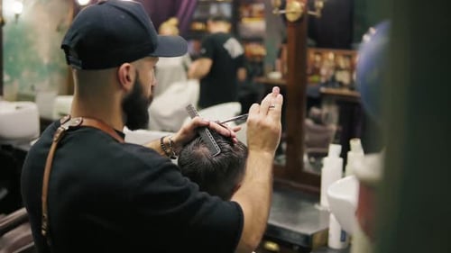 Vista posterior del elegante barbero barbudo con una gorra negra con un delantal de cuero realizando un corte de pelo
