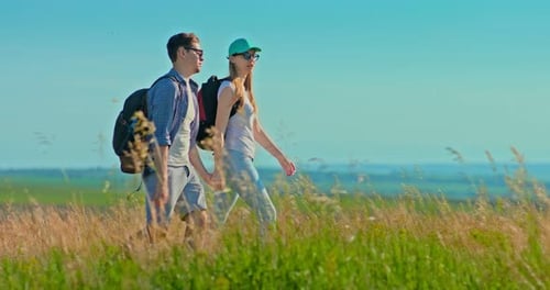 Pair of Lovers is Walking Across the Field with Backpacks and Holding Hands