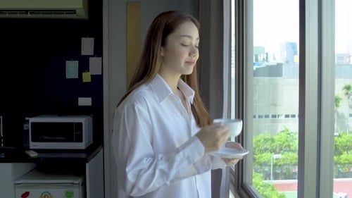 Side view of mixed race woman in pajamas standing by window with cup of tea or coffee