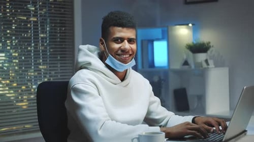 Bel homme noir avec masque médical souriant à la caméra tout en travaillant à la maison
