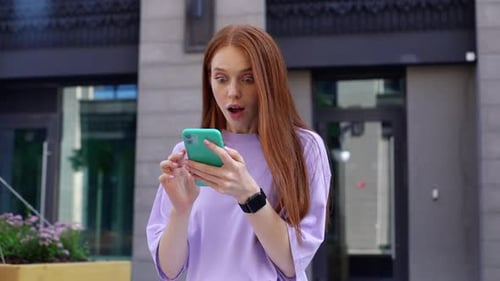 Surprised Redhead Lady Reading Good News on Phone Outdoors Background of City Street.