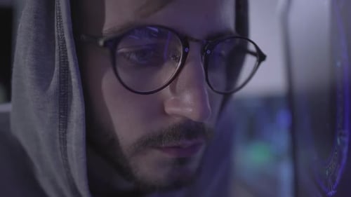 Nahaufnahme eines bärtigen jungen Mannes mit Brille und Kapuze im Innenbereich. Männlicher kaukasischer Hacker oder Programmierer