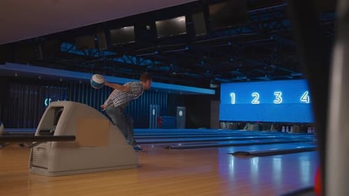 In einem karierten Hemd wirft ein Mann in einer Bowlingbahn einen Ball in Zeitlupe und schlägt Kegeln nieder