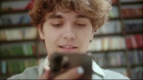 Portrait of Happy Young Student Chatting By Mobile Phone in University Library