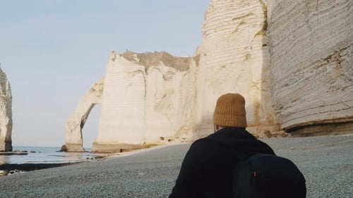 Rear View Caucasian Tourist Man with Backpack Walking Along Sea Shore To White Arch Cliff at Sunny
