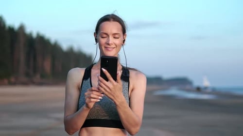 Smiling Fitness Woman Listening Music in Earphones Use Smartphone at Sunset Beach Panning Shot