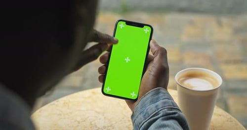 Hände eines afroamerikanischen Mannes, der am Tisch sitzt, mit Kaffeetasse aus Papier darauf und Smartphone mit