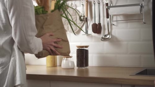 Mujer pone una bolsa de papel ecológica con verduras frescas y baguette sobre la mesa de una cocina moderna