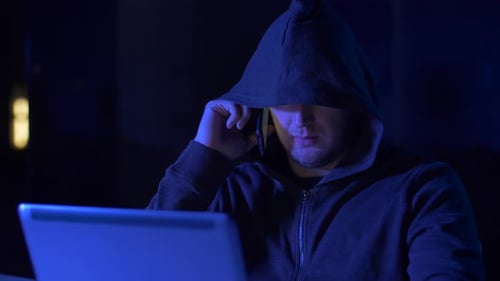 Hooded Hacker Using Laptop and Talking on Phone