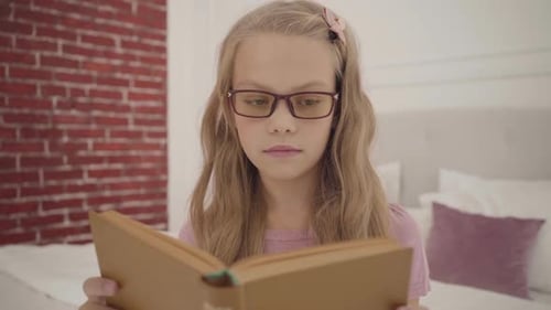 Young Intelligent Teenage Girl in Glasses Reading the Book at Home