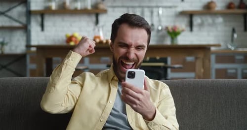 Man Celebrates Success While Using Mobile Phone on Sofa