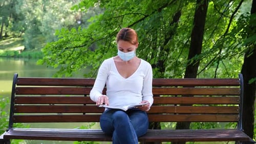 Young Woman Sits Alone in a Park in a Mask and Reads a Book During the Epidemic