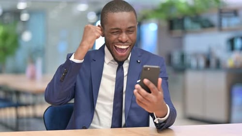 African Businessman Celebrating Success on Smartphone in Office