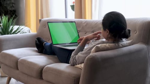 Young Woman at Home Works on a Laptop Computer with Green Mock-up Screen