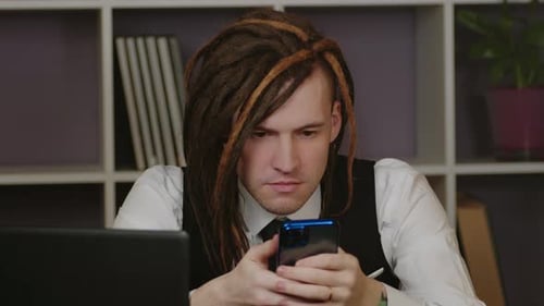 Young Man Sitting at Laptop Using Smartphone in Home Office