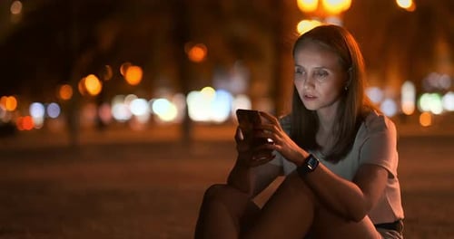 A Young Girl on the Background of the Night City Looks Into the Smartphone Screen