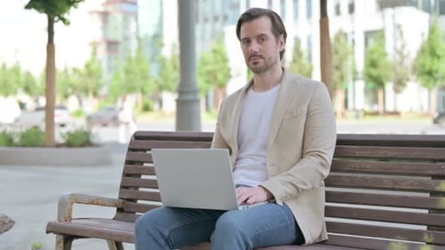 Man Using Laptop on Bench in Urban Park