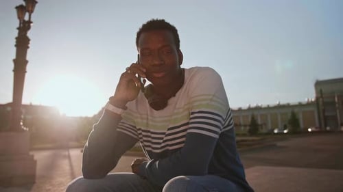 Ein junger schwarzer Student in stylischer Kleidung sitzt und spricht auf einem Smartphone in der Stadt