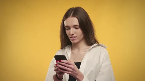 Young Adult Caucasian Woman is Laughing with a Smartphone in Hands on a Bright Yellow Background