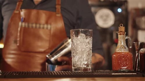 Bar Rack with Tall Glass with Ice Cubes Inside Barman Is Moving in Background