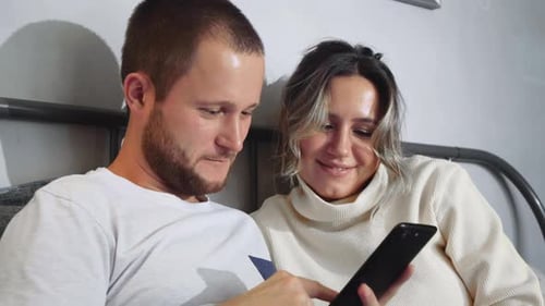 A happy young couple are laying in the bed at home while looking at the mobile phone together
