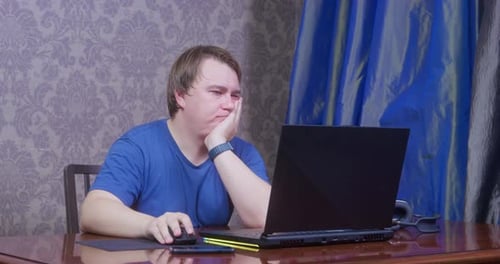 Junger Mann im blauen T-Shirt macht sehr langweilige und eintönige Arbeit im Home-Office mit Laptop, damit er