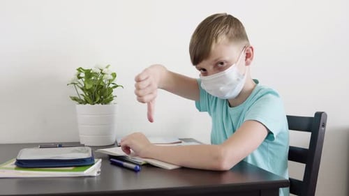 A Young Boy in a Face Mask Shows a Thumb Down To the Camera and Shakes His Head As He Sits at Table