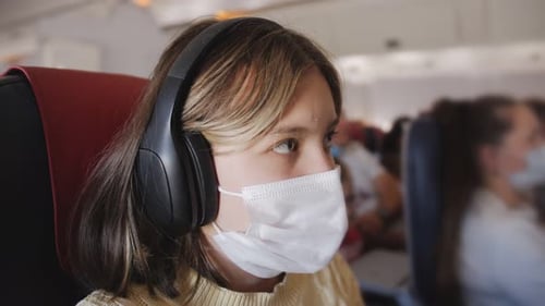 A Teenage Girl in the Cabin of an Airplane in a Protective Mask Listens to Music with Headphones