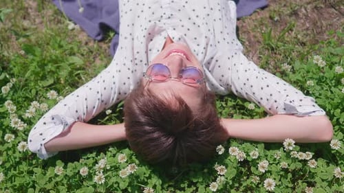 Blick von oben auf Frau mit bunter Sonnenbrille, die auf Rasen im Stadtpark liegt und Blumen genießt