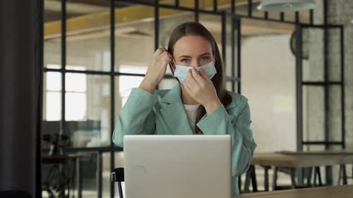 Young Woman Puts a Medical Mask on Her Face and Works at a Laptop in the Office