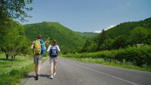 A Couple of Tourists with Backpacks are Walking Along the Asphalt Road Towards the Beautiful Green