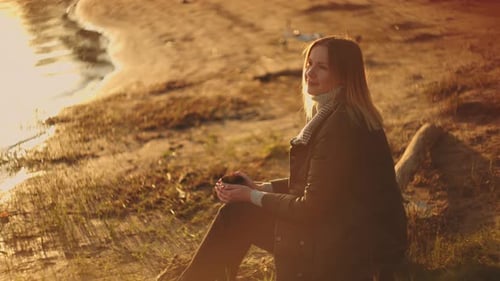 A Beautiful Young Woman Enjoys a Mug of Fragrant Drink in the Sun's Rays Watching the Sunset and on