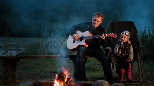 Man Plays Guitar With Child Next to Campfire