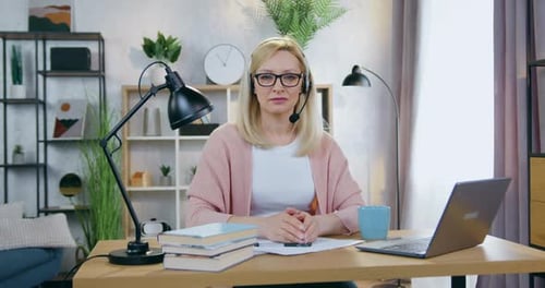Mujer con auriculares y anteojos mirando a la cámara en la oficina en casa durante una videoconferencia