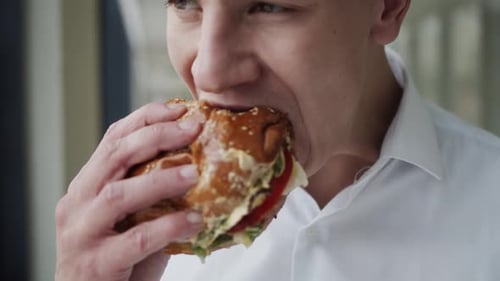 Close View of Handsome Man in Shirt Eating Tasty Hamburger Indoor at Window