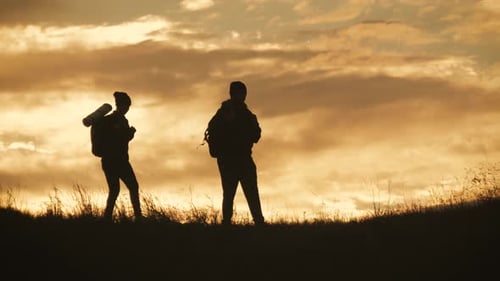 Silhouettes of Two Hikers with Backpacks Enjoying Sunset View From Top of a Mountain