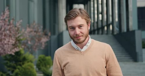 Retrato de un hombre guapo de éxito, mirando hacia arriba a la cámara, sonriendo al fondo del edificio de oficinas.